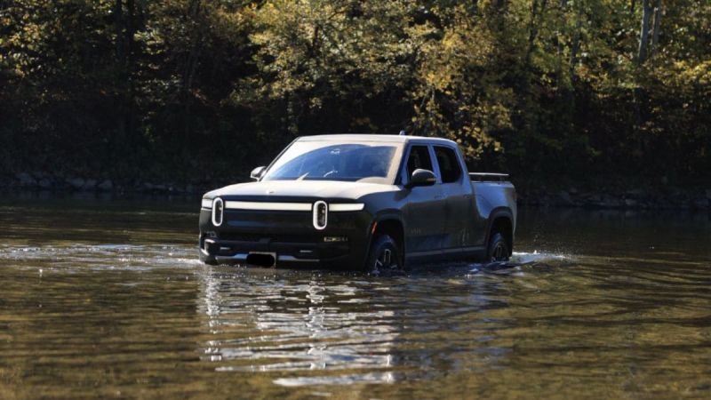 Rivian R1T electric pickup truck crossing a river, surrounded by autumn forest with water reflecting its metallic blue-gray body
