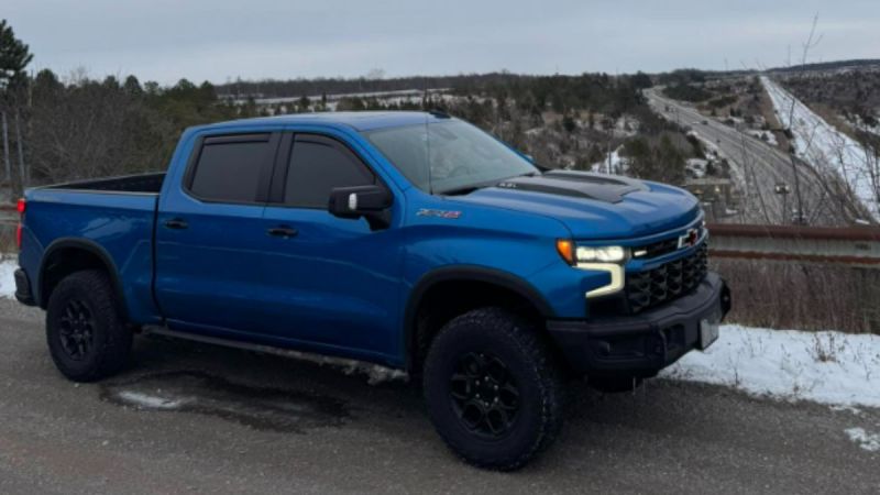 Bright blue Chevrolet Silverado ZR2 pickup truck with black wheels parked on snowy roadside with highway backdrop