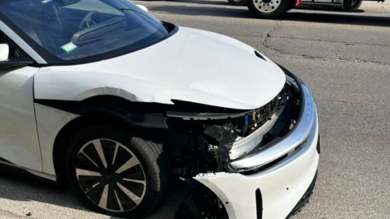 A white Lucid Air Touring with significant front-end damage, including a missing bumper and exposed internal components, is parked on a city street during daylight.