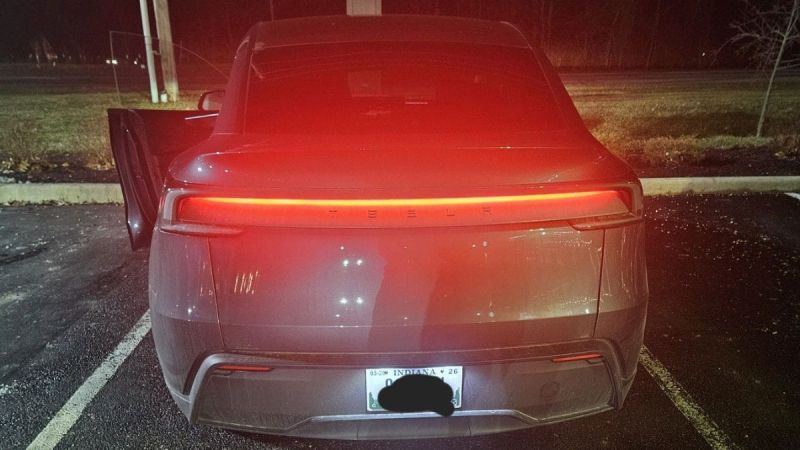Rear view of a Tesla Model Y parked at night with the brake lights glowing red. The driver's door is slightly open, and the surroundings are dimly lit.