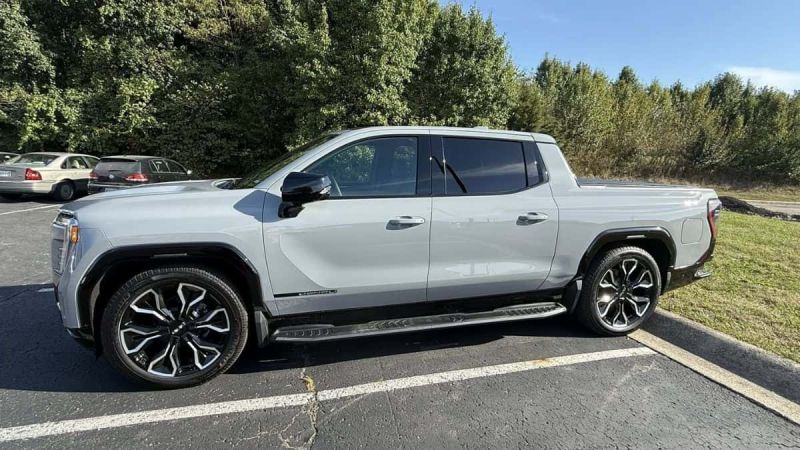 A grey GMC Sierra EV with large and stylish wheels is parked on a paved area. Surrounded by green trees, the scene conveys a modern, outdoorsy vibe.
