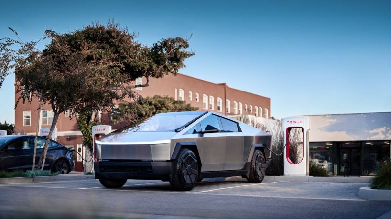 A Tesla Cybertruck parked at a charging station near a modern building, with trees and another Tesla vehicle in the background.