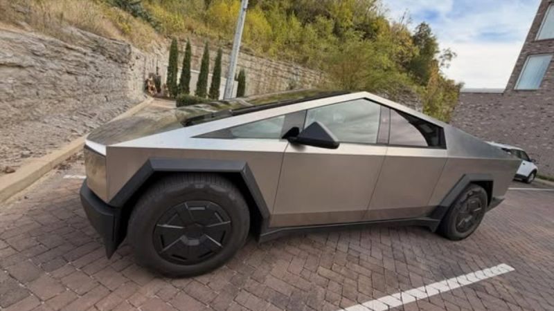 The futuristic and angular Tesla Cybertruck in a parking lot surrounded by a stone wall and greenery. The design is sleek and modern with a bold presence.