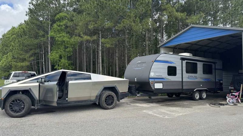 A 2025 Tesla Cybertruck parked next to a gray travel trailer under a metal shelter, surrounded by dense green trees.