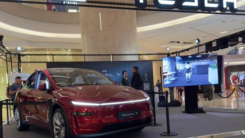 A red Tesla Model Y is showcased in a modern mall setting, with a digital screen displaying a robotic character beside it.