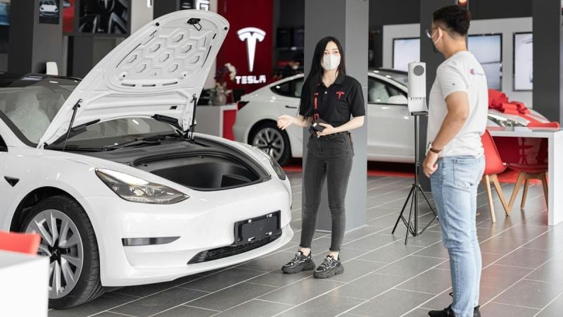 A Tesla sales representative explains features of a white Tesla Model 3 with the hood open, in a sleek showroom setting.