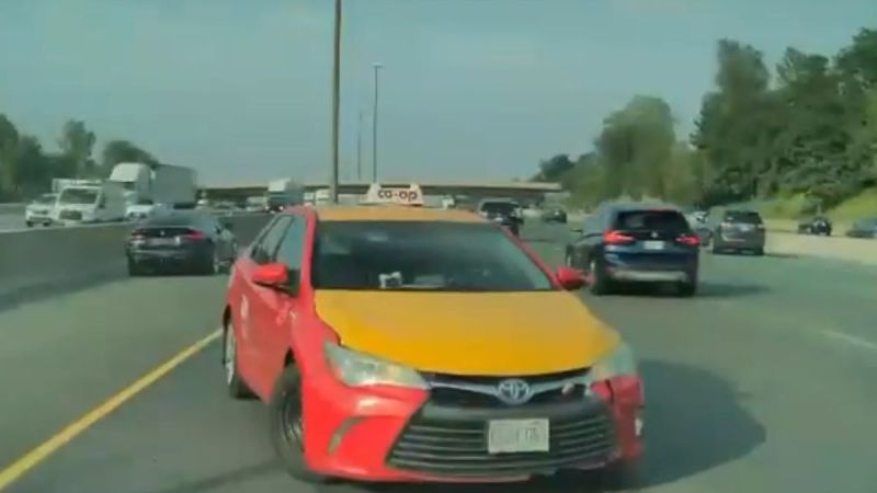 A red and yellow Toyota taxi labeled "co-op" is spun out on a multi-lane highway. Surrounding cars and lush trees line the road under a clear sky.