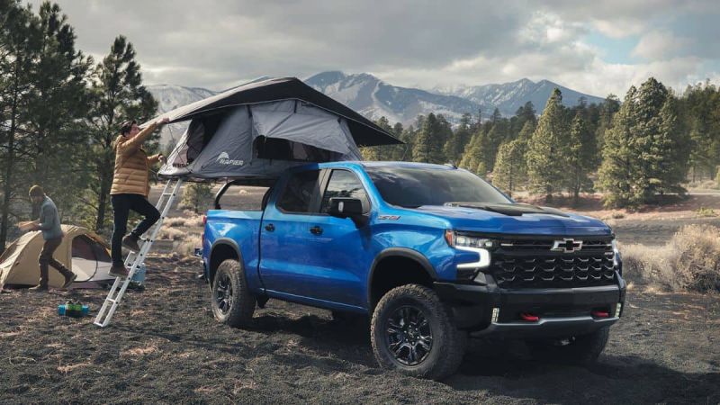 A blue Chevrolet Silverado 1500 truck with roof tent in a forested mountain area. A person climbs the tent ladder and another person assembles a ground tent nearby. Atmosphere is a peaceful outdoor scene.
