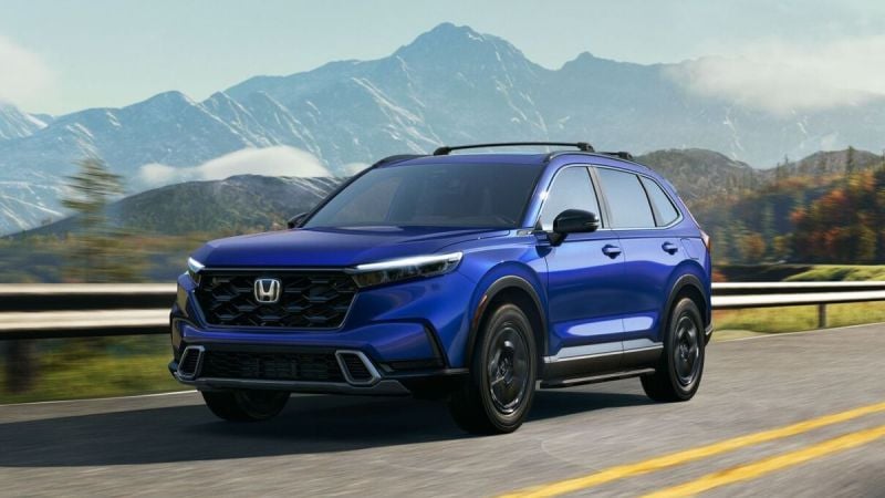 A blue 2025 Honda CR-V drives on a highway with mountains in the background under a clear sky. The scene conveys a sense of adventure and freedom.