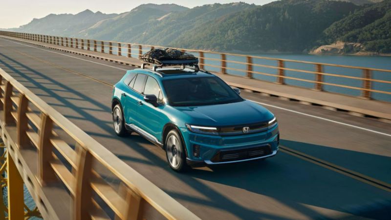A turquoise 2026 Honda Prologue is shown from a three-quarter front angle driving across a bridge with mountains and a lake in the background, featuring a roof-mounted cargo carrier.