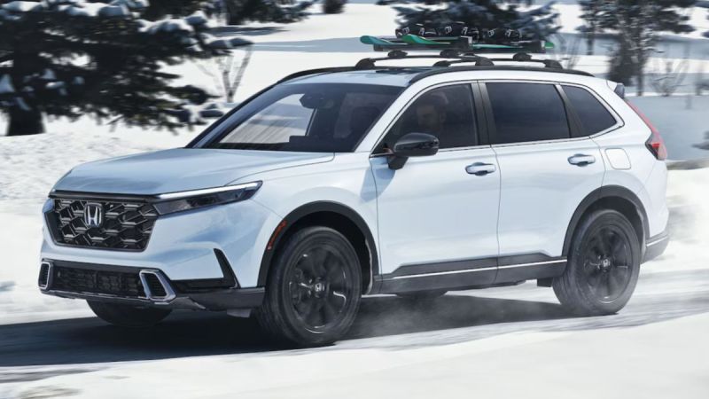 A white Honda CR-V drives on a snow-covered road with trees in the background. It has a roof rack carrying skis, conveying a sense of adventure and winter travel.