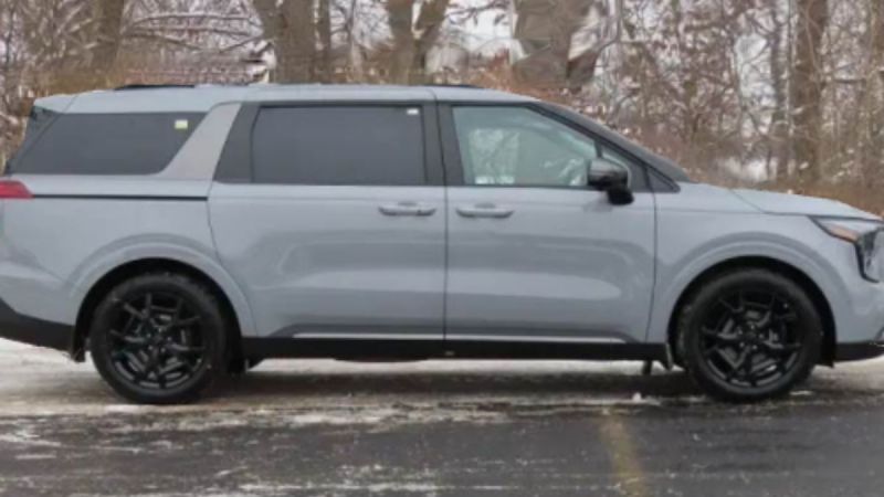 A grey Kia Carnival is shown from the side view, featuring black wheels and trim, photographed in a winter setting with snow-covered trees in the background.