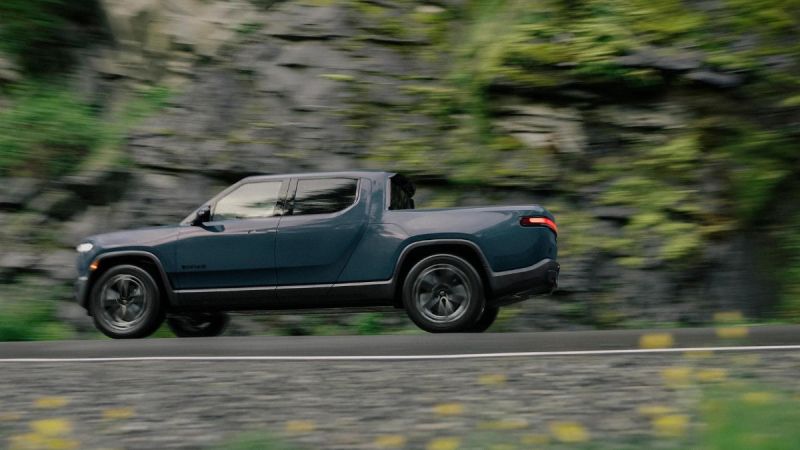 Dark blue electric pickup truck speeding along rocky mountain road with blurred green forest background