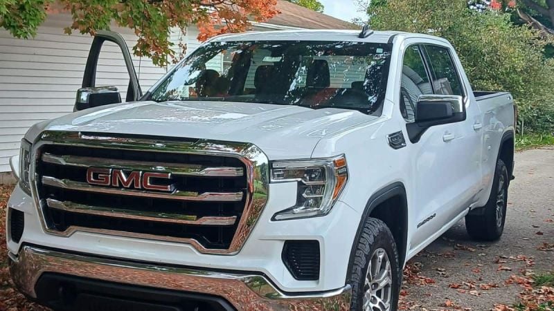 White 2021 GMC 1500 parked in a driveway surrounded by fall foliage. The driver's door is open, conveying readiness and adventure.