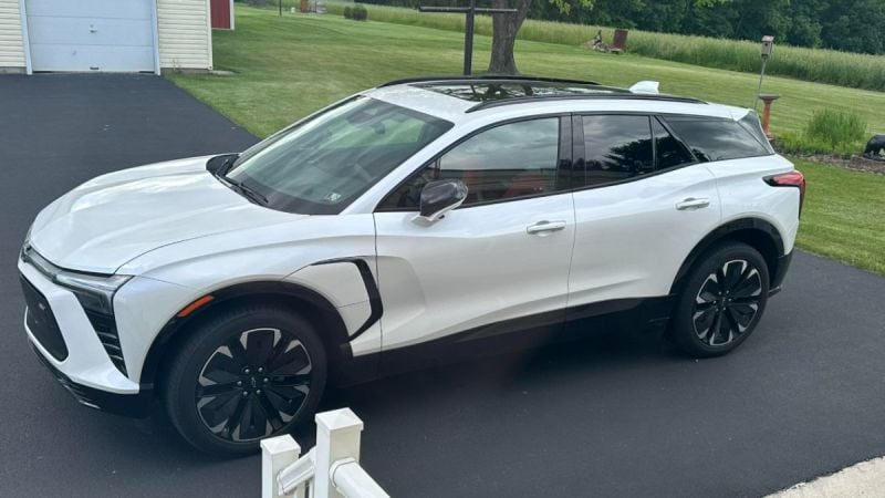 White Chevrolet Blazer EV SUV parked on black asphalt driveway with green lawn and trees in background, sleek modern design with black wheels