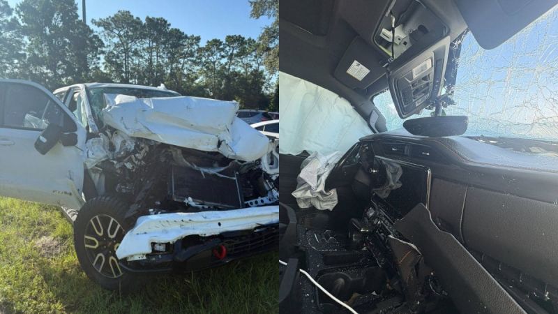 A totaled white GMC Sierra 1500 AT4 is shown in a grassy area, its front crumpled from a collision. The interior displays deployed airbags and a cracked windshield.
