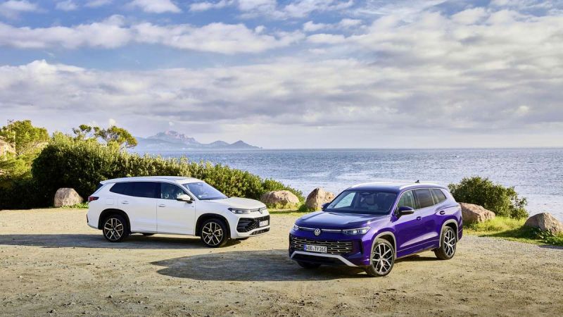 Two modern 2025 Volkswagen Tayrons, a white one and a purple one, parked by the seaside with a scenic view of mountains and clouds in the background.