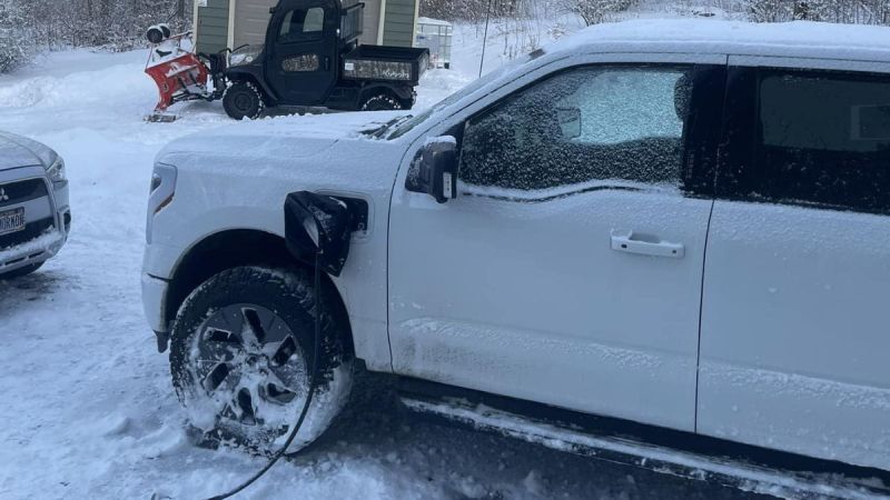 White Ford F-150 Lightning pickup truck shown from the side angle in snowy winter conditions, with snow-covered wheels and a utility vehicle with plow attachment visible in the background.