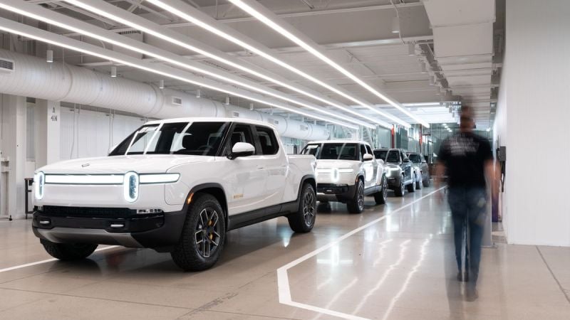 A row of white Rivian R1T in a modern workshop, with a blurred figure walking past under bright overhead lighting.