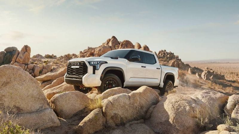 A white 2025 Toyota Tundra truck navigates rocky terrain under a clear sky, showcasing its rugged design and off-road capability.