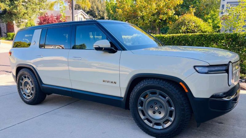 A Rivian electric SUV parked in a driveway, showcasing its sleek design, rugged tires, and modern details against a green backdrop.