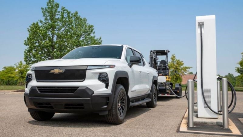 A white Chevrolet truck is parked beside an electric charging station, with green trees and a clear blue sky in the background.