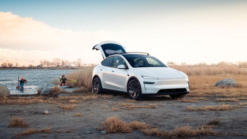 A white 2026 Tesla Model Y Juniper parked by a lake, with two people kayaking nearby under a colorful sky and tall grass.