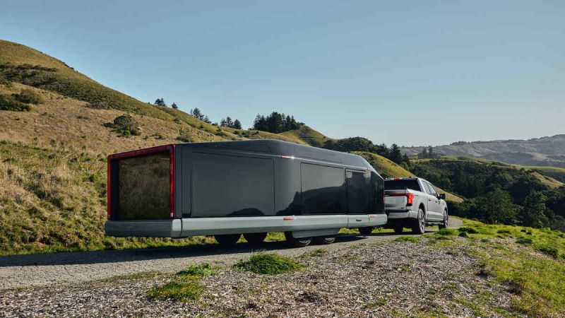 Image of the Lightship L1 camper being towed behind a Ford F150 on a dirt road with green rolling hills surrounding.