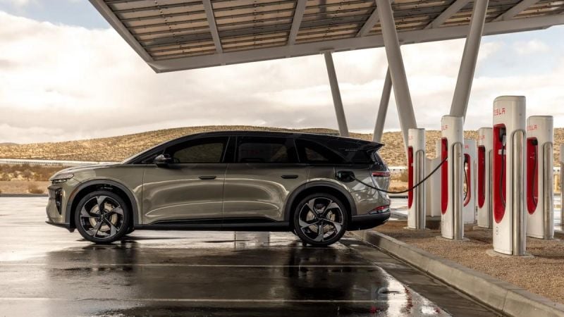 Lucid Gravity electric SUV charging at a Tesla Supercharger station, side profile with multiple charging stalls in a desert landscape.
