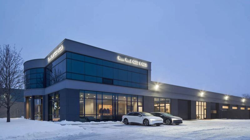 Image showing a pair of Lucid Airs parked outside Lucid's huge new combination Studio/Service Centre in West Montreal. The building looms large against the snow and steely grey sky, with the Lucid logo illuminated on two sides of the facade.
