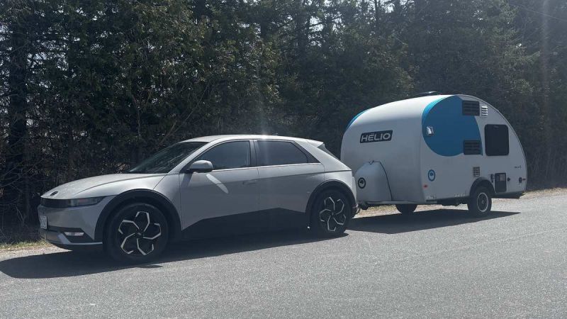 A 2025 Hyundai Ioniq 5 towing a compact, modern camper trailer on a quiet road, framed by tall green trees.