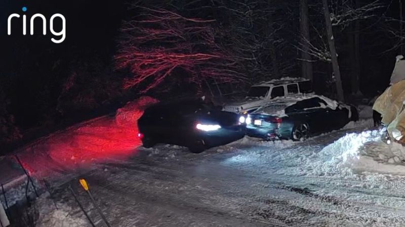 Ring camera footage at night showing a dark Ford Mustang Mach-E with bright headlights and red taillights, and a dark sedan, stuck or maneuvering in a snow-covered driveway.