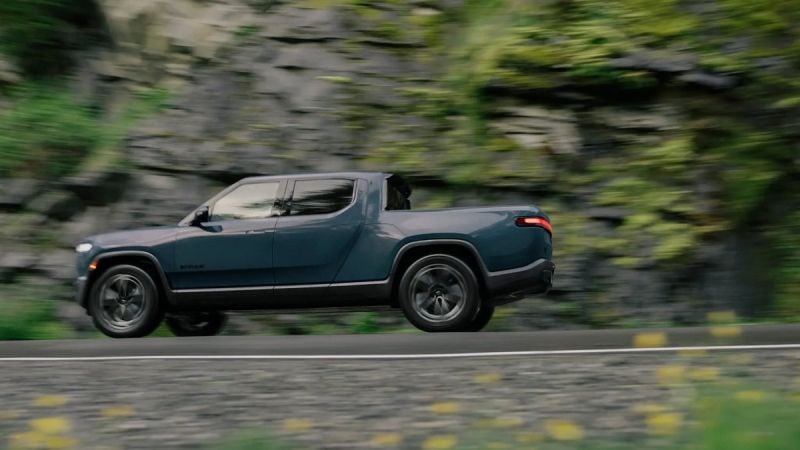 Side view of a Rivian R1T electric truck driving on a road with a rocky, green hillside background, motion blur showing speed.