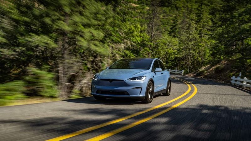 Tesla Model X driving on a winding road through forest landscape, side profile view