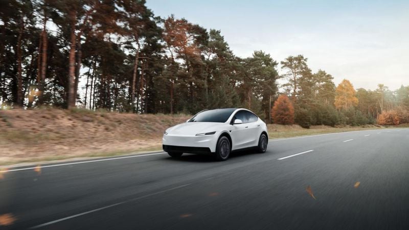 White Tesla Model Y electric SUV driving on scenic highway with forest and autumn trees
