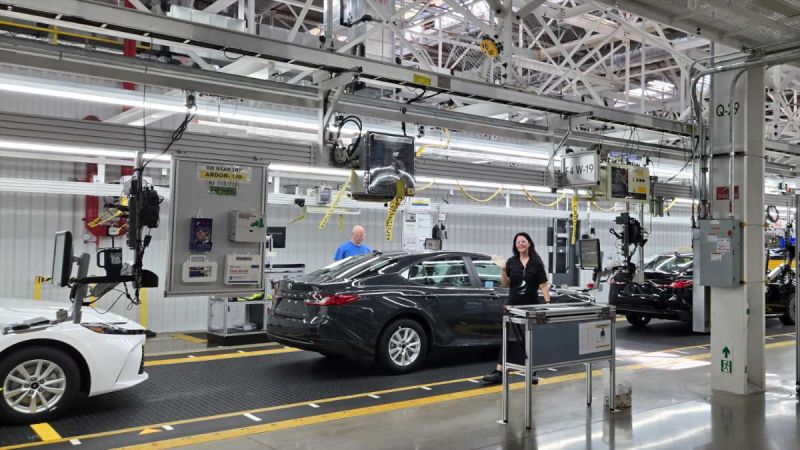 A Toyota factory line worker waves hello while building an American-made Camry on an assembly line