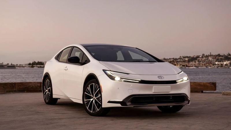 White Toyota Prius hybrid sedan parked near the water at golden hour, angled front view highlighting sleek exterior lines.