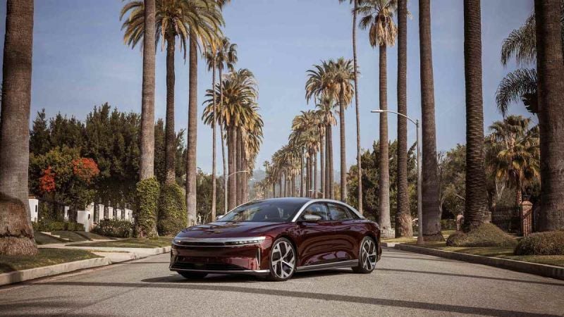 A Lucid Air in Zenith Red is pictured parked in the center of a sun-soaked Californian street lined with tall palm trees. Mountains are visible in the background.