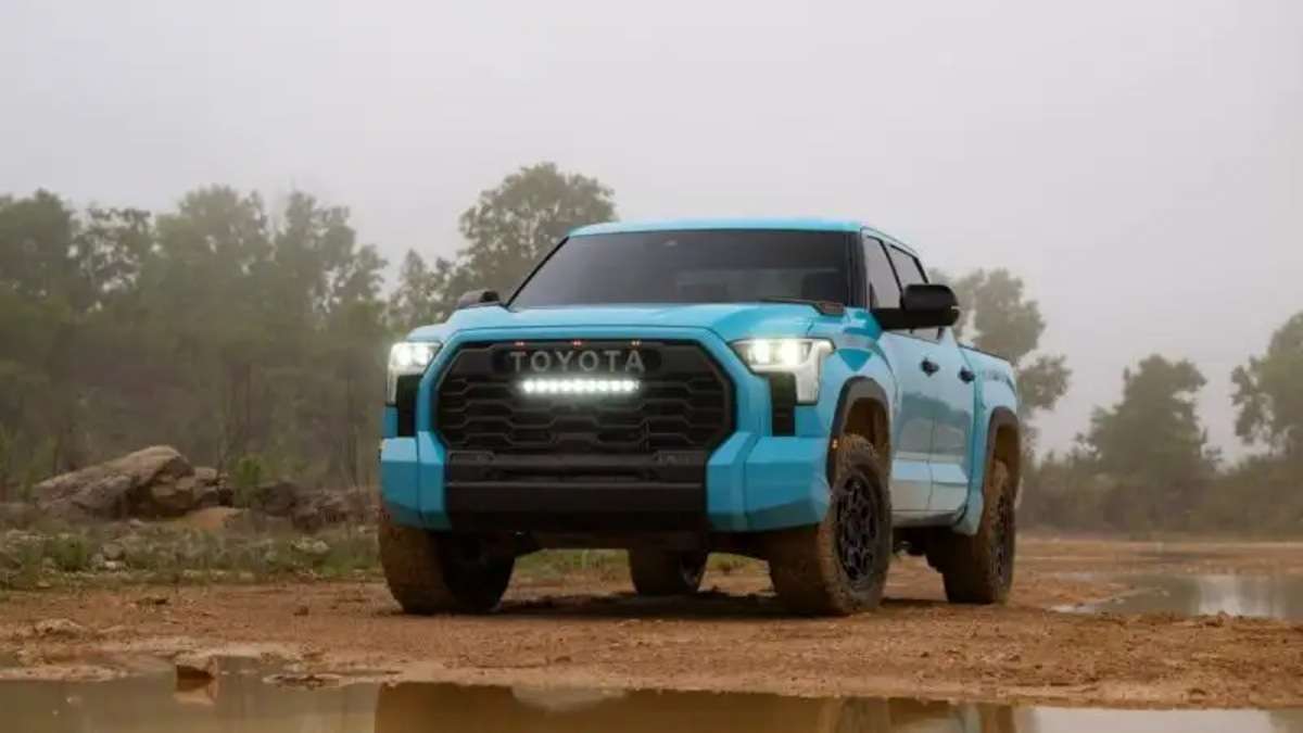 A head-on shot of a light blue 2025 Toyota Tundra TRD Pro with its distinctive LED light bar turned on, parked in a muddy, off-road environment.