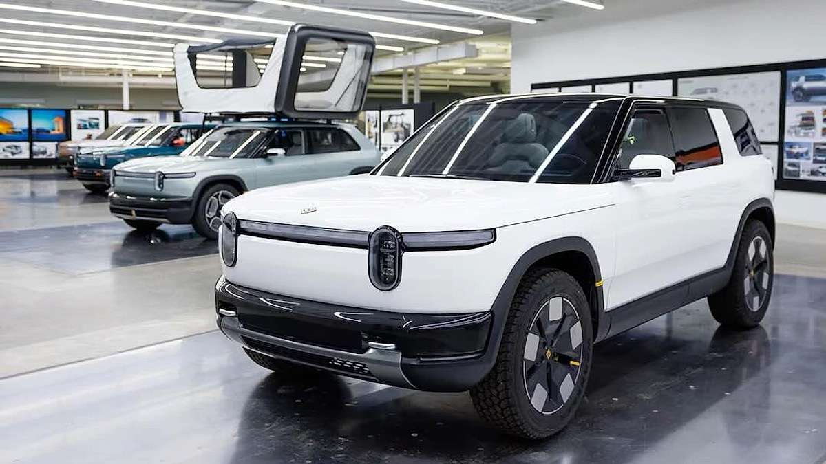 White Rivian R2 compact electric SUV concept on display in Rivian design studio alongside teal R2 and R1S prototypes with design sketches on wall in background