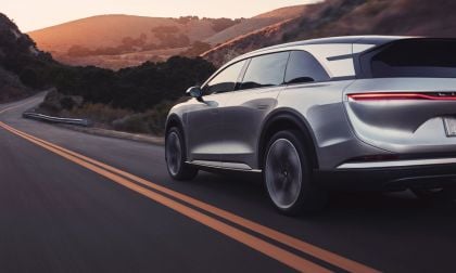 A sleek silver Lucid Gravity SUV drives along a winding road at sunset, surrounded by hills and lush greenery.