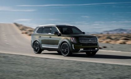 A dark gray/olive 2020 Kia Telluride SUV shown from a front three-quarter angle, driving on a mountain road with black wheels and trim accents against a desert landscape and blue sky.