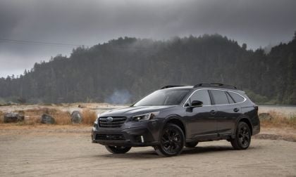 A dark gray 2021 Subaru Outback is shown from a front three-quarter view, parked on a gravel area with misty mountains and forest in the background.
