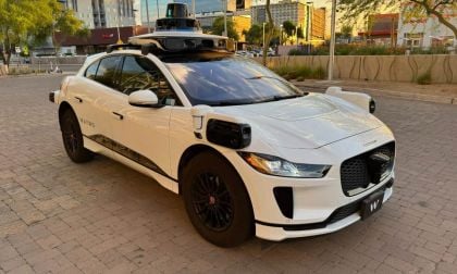 White Jaguar i-Pace with Waymo autonomous driving sensors, parked on brick pavement with urban backdrop