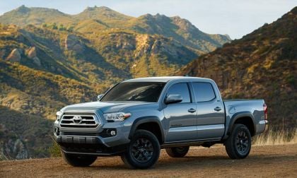 Front three-quarter view of a silver Toyota Tacoma off-road truck parked on a mountain overlook with rugged terrain behind it.