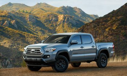 Front three-quarter view of a silver Toyota Tacoma off-road truck parked on a mountain overlook with rugged terrain behind it.