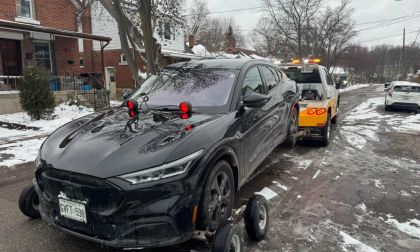 A black Ford Mustang Mach-E electric SUV, likely a 2022 model, is shown being loaded onto a tow truck from its front three-quarter angle on a snowy residential street.