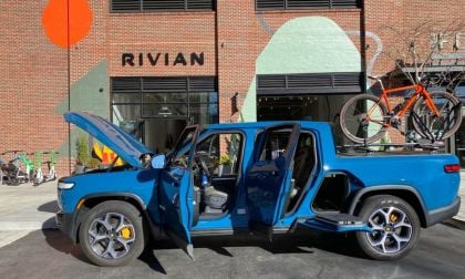 Electric blue Rivian R1T pickup truck with open doors, parked in front of Rivian headquarters, with orange bicycle mounted on roof rack.