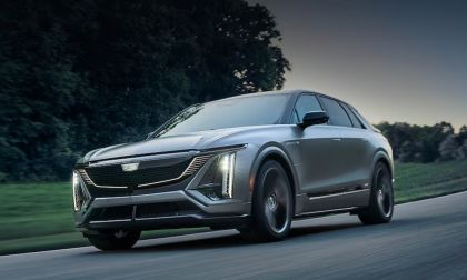 2023 Cadillac Lyriq electric SUV driving on a road, front three-quarter view showing illuminated LED grille and vertical headlights.