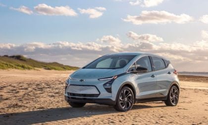 A silver Chevrolet Bolt EV is parked on a sandy beach under a clear blue sky with scattered clouds. The scene conveys a serene and eco-friendly vibe.